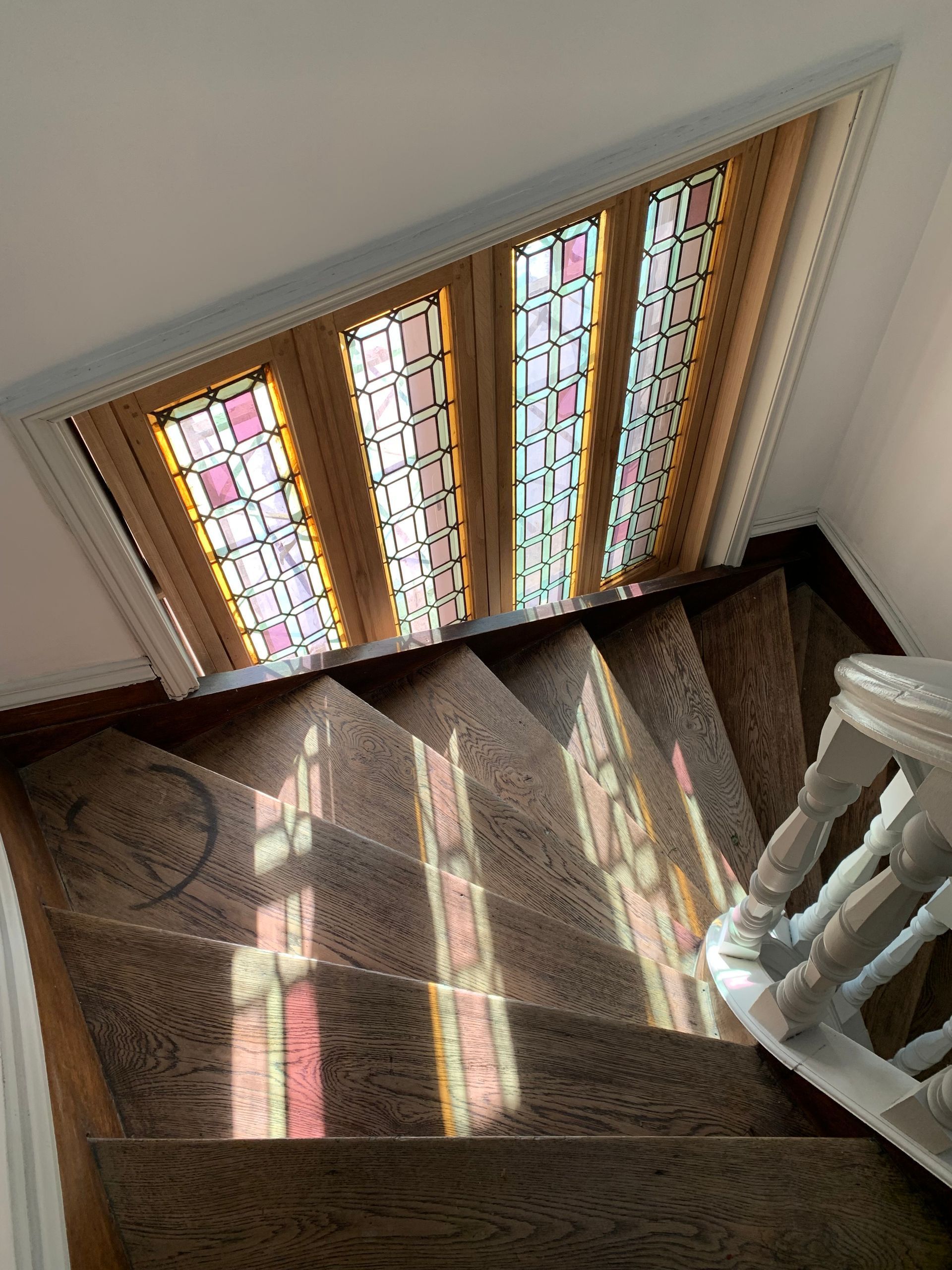 Palier d'escalier baigné de soleil, orné de vitraux projetant une lumière colorée sur du parquet et une rampe en bois.