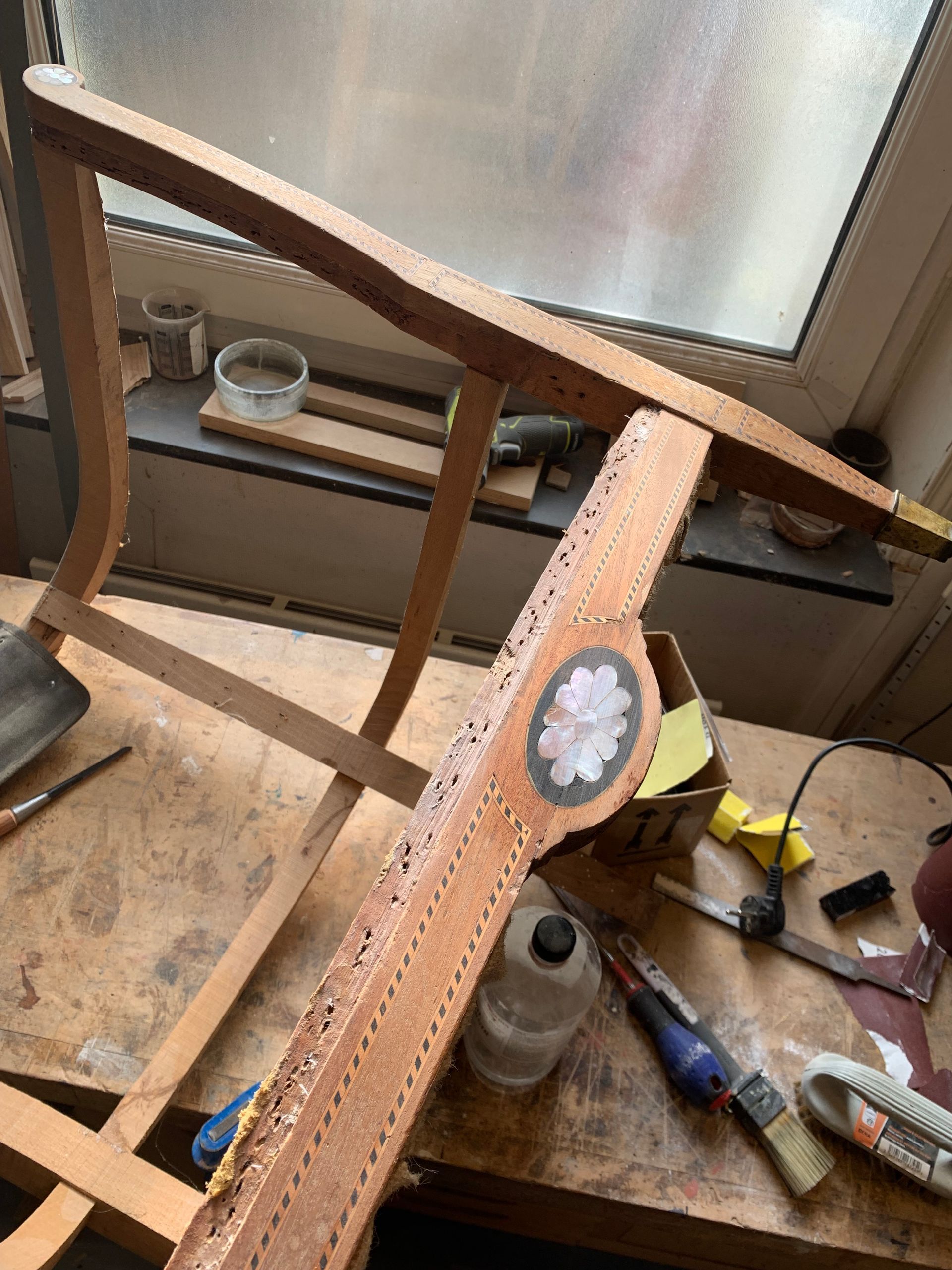 Chaise en bois en cours de réparation sur un établi, avec des outils et une fenêtre dépolie en arrière-plan.