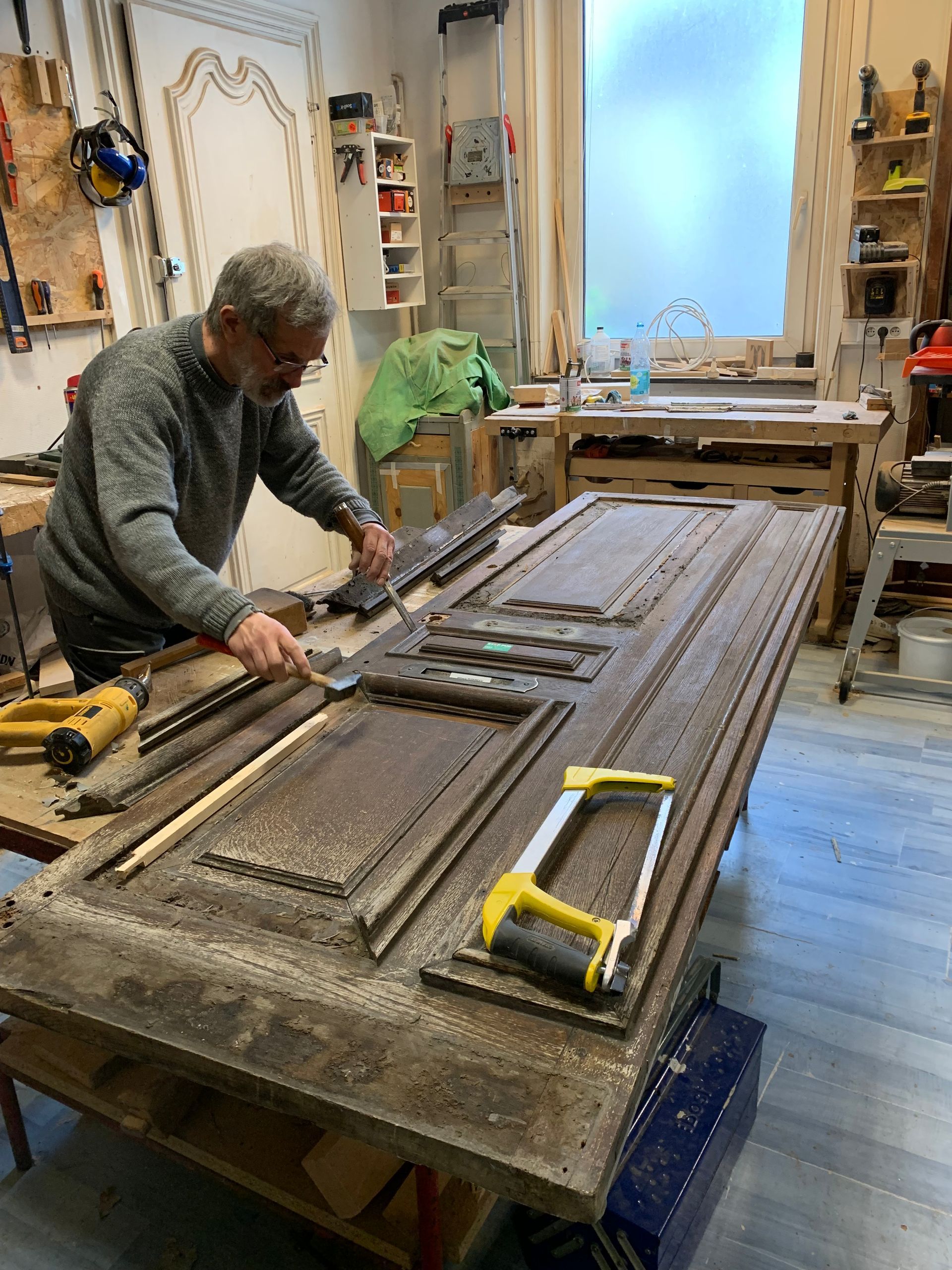 Une personne travaille sur un grand panneau de bois dans un atelier, à l'aide de serre-joints et d'outils manuels.