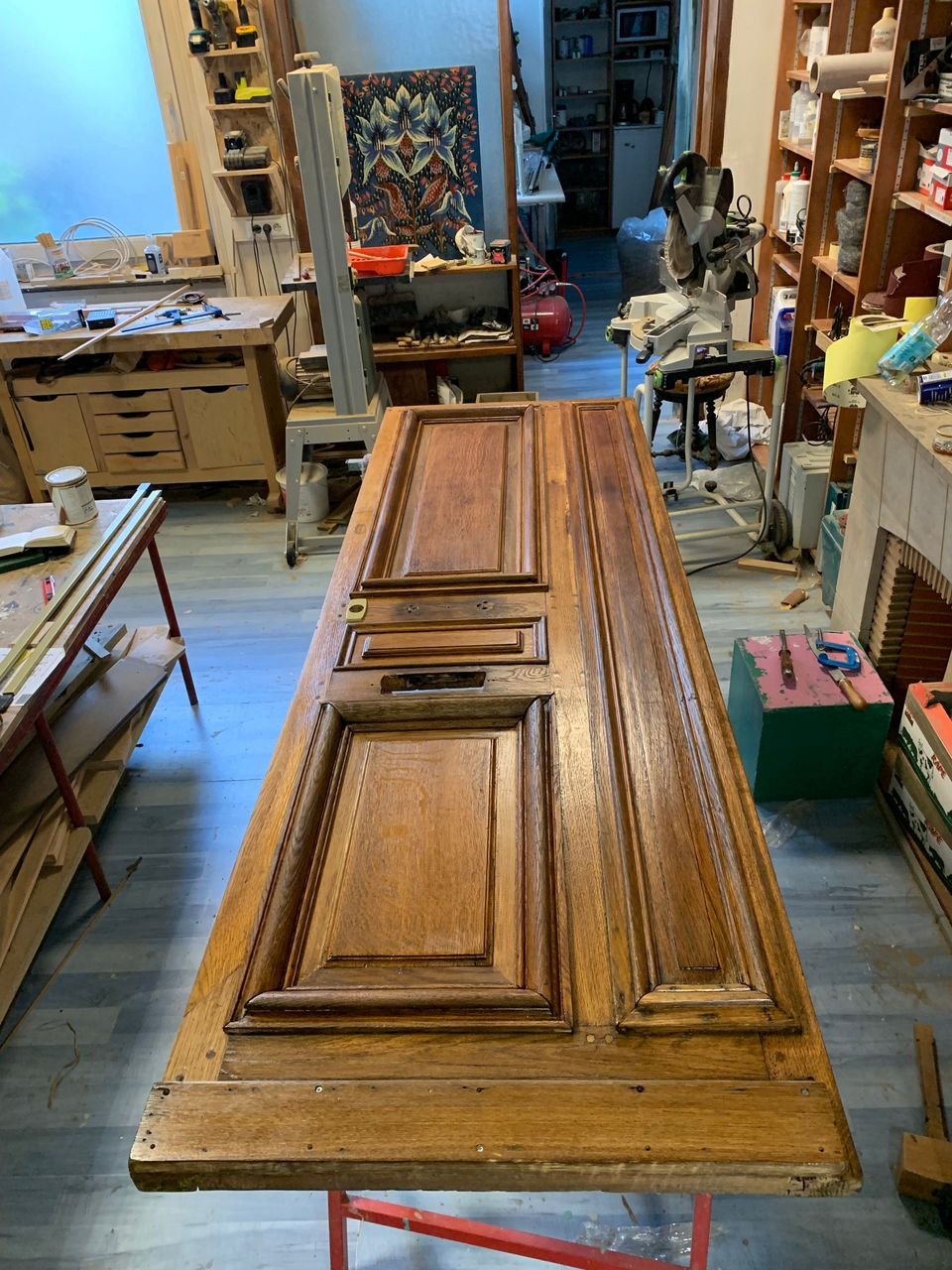 Une porte en bois à panneaux, posée à plat dans un atelier encombré d'outils et de meubles.