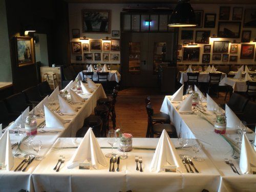 Restaurantinterieur mit langen, für eine Mahlzeit gedeckten Tischen, weißen Tischdecken, gefalteten Servietten und Kunstwerken an den Wänden.