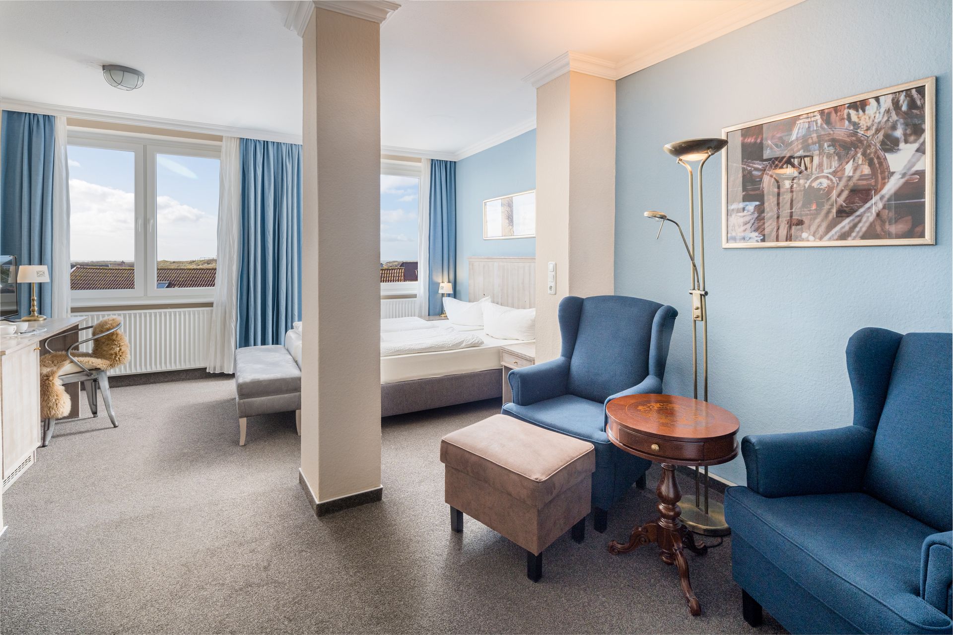 Hotelzimmer mit Bett, Stühlen und Fenstern mit blauen Vorhängen.