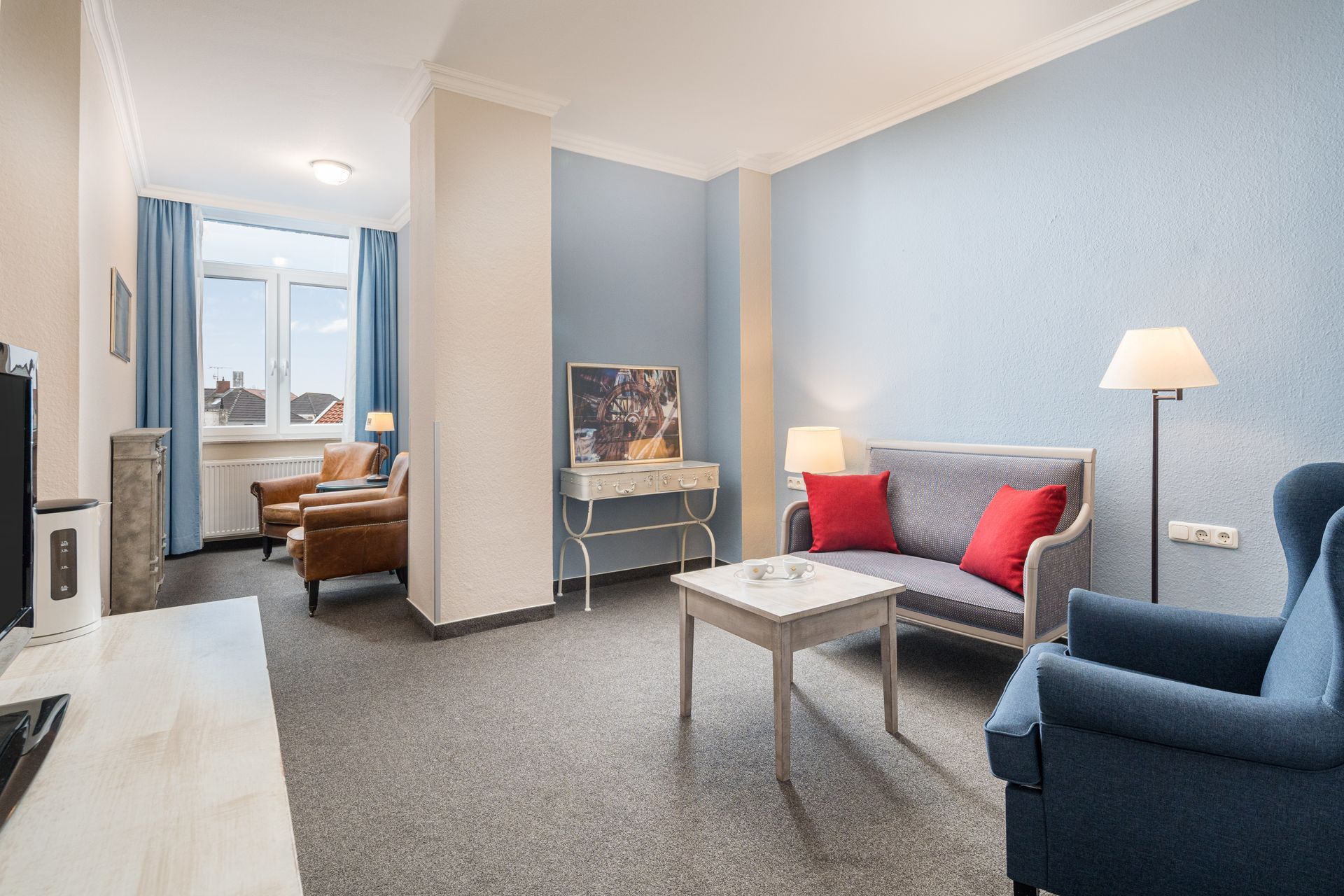 Wohnzimmer mit blauen Wänden, grauer Couch und Sesseln, Couchtisch und Fenster mit blauen Vorhängen.