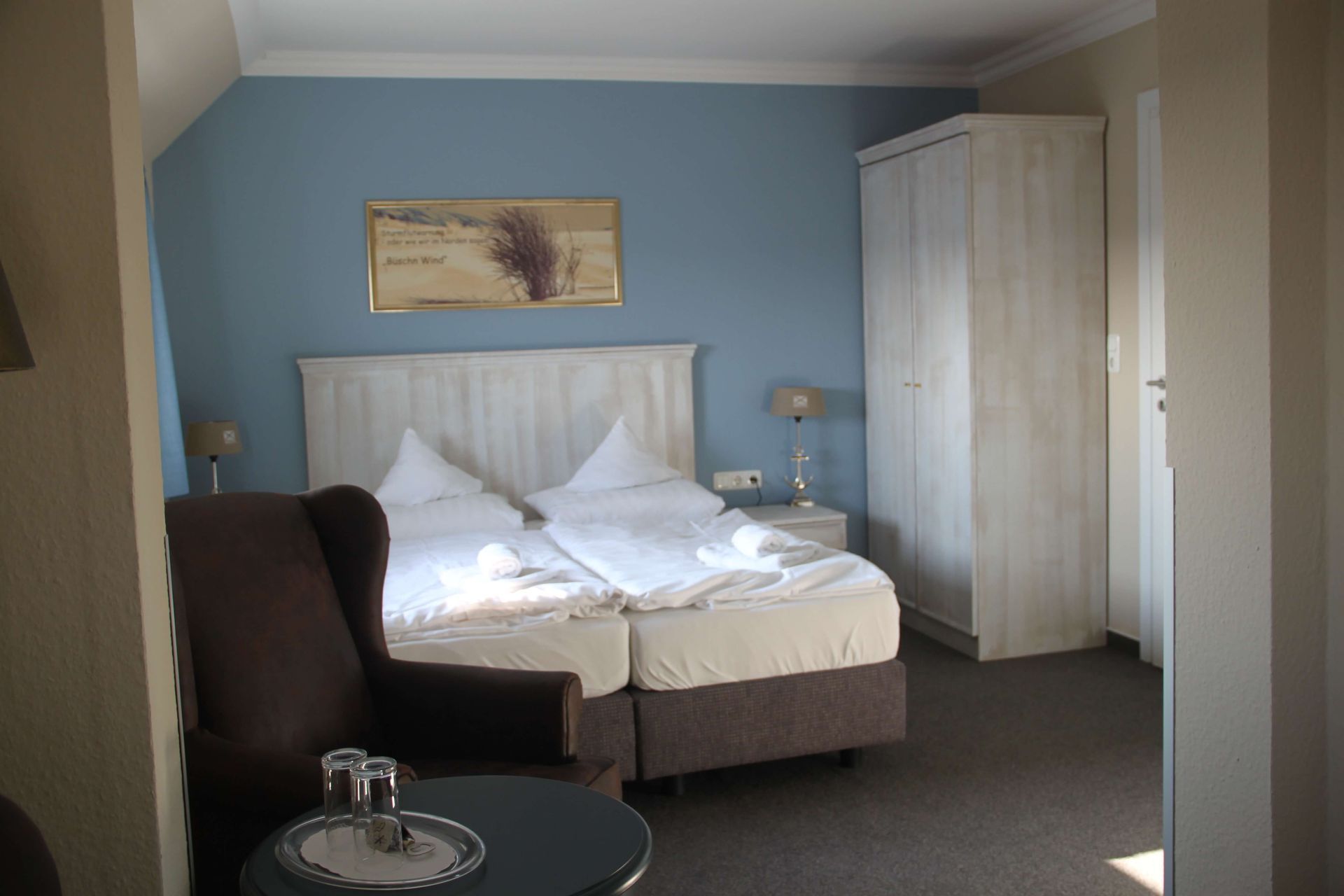 Schlafzimmer mit Bett, Kleiderschrank, Sessel, blauer Wand und Kunstwerken.