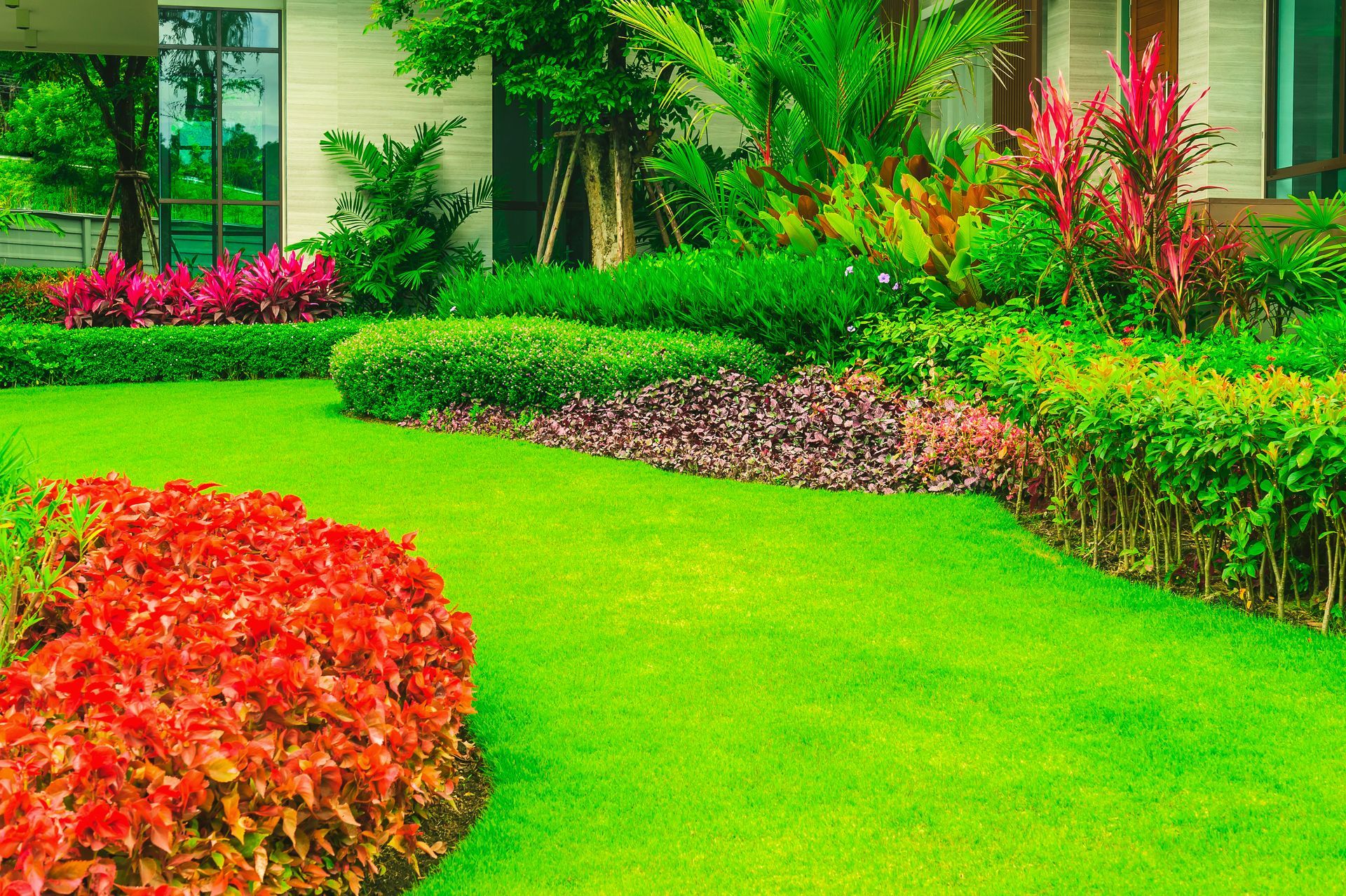 Un jardin luxuriant avec une pelouse d'un vert éclatant, des plantes ornementales rouges, des haies taillées et des arbres denses près d'un bâtiment.