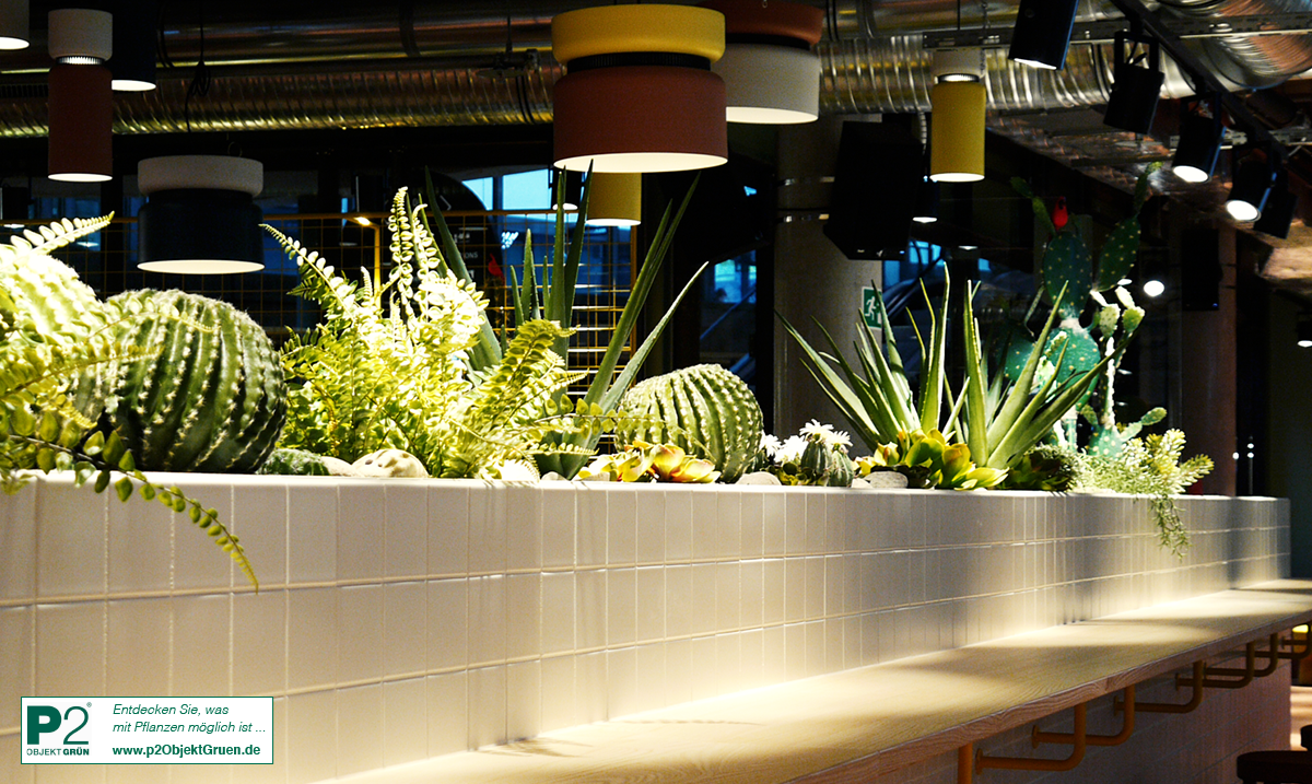Beleuchteter Innenraum mit grünen Pflanzen und weißen Kacheln an einer langen Theke