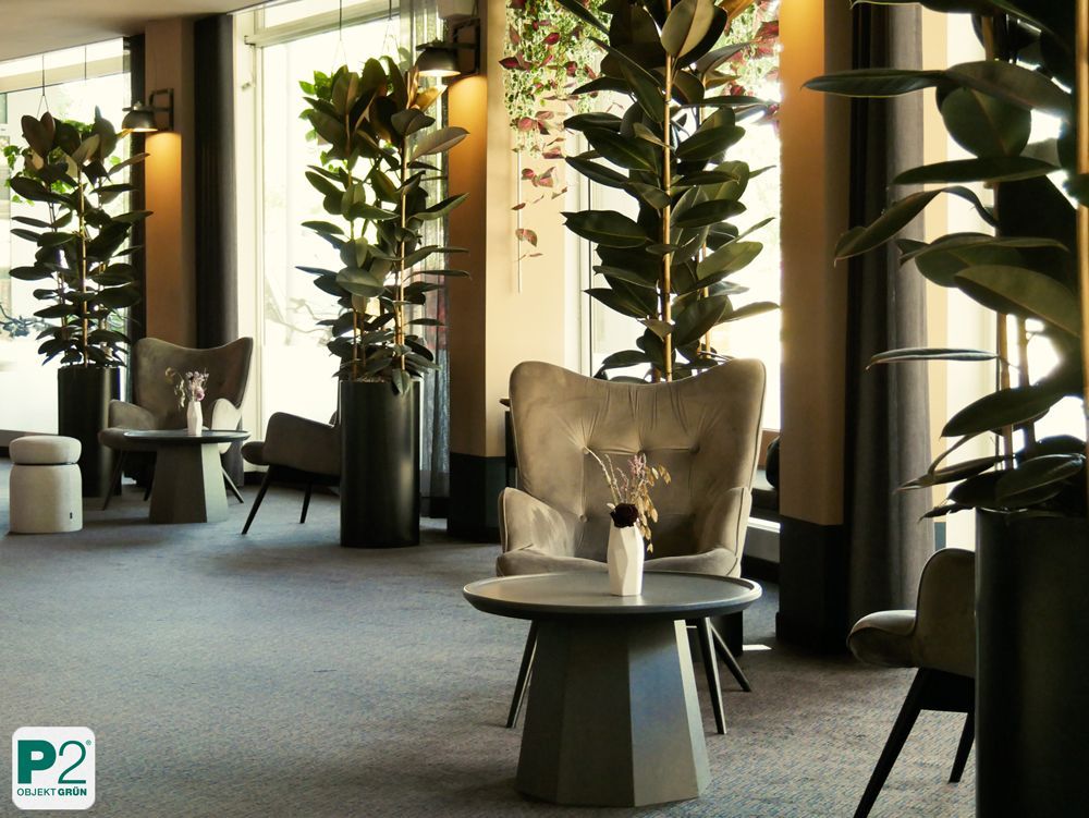 Gemütliche Sitzecke mit grauen Sesseln, runden Tischen und großen Zimmerpflanzen in einem hellen Raum