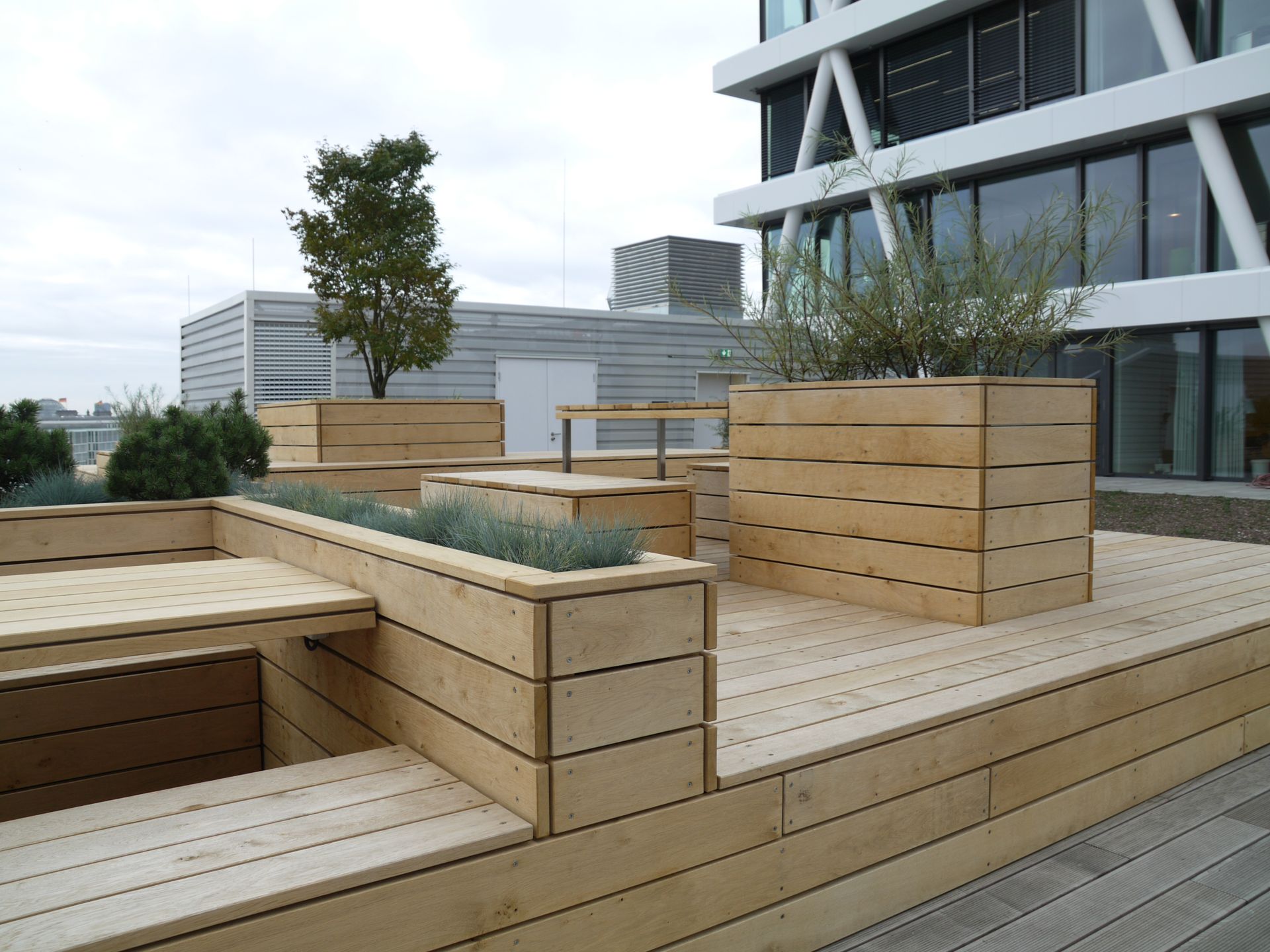 Eine moderne Dachterrasse mit Holzbänken, Pflanzkübeln mit Grünpflanzen und kantigen Holzbelägen.