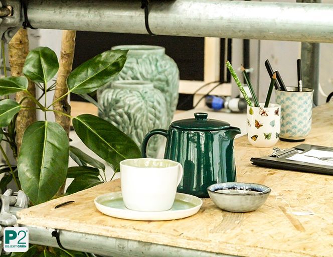 Weißer Kaffeebecher auf Untertasse neben grünem Teekännchen und Stiftehaltern auf Holzregal mit Pflanzen im Hintergrund