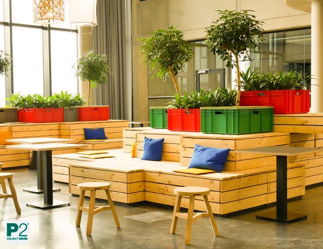 Heller Arbeitsbereich mit Holzmöbeln, bunten Kissen und großen Pflanzkübeln als Raumteiler.