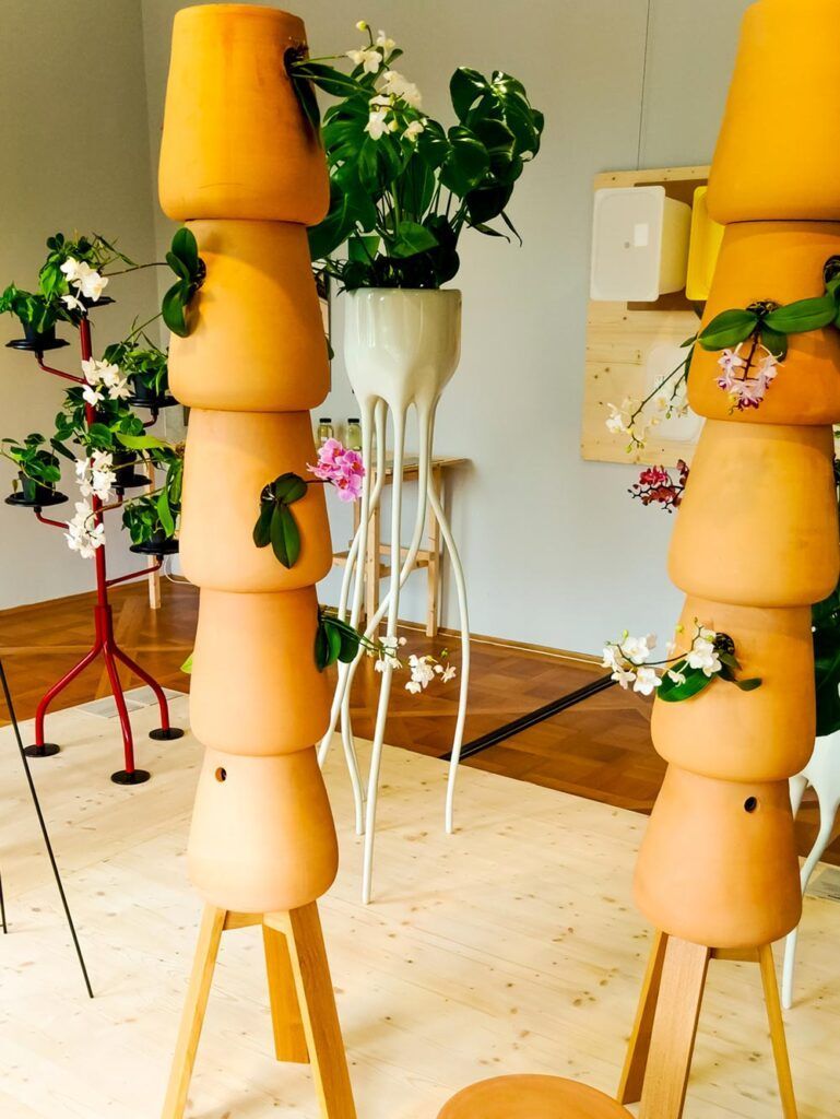 Organisch geformte Pflanzgefäße mit Blüten auf Holzplattformen im Ausstellungsraum.