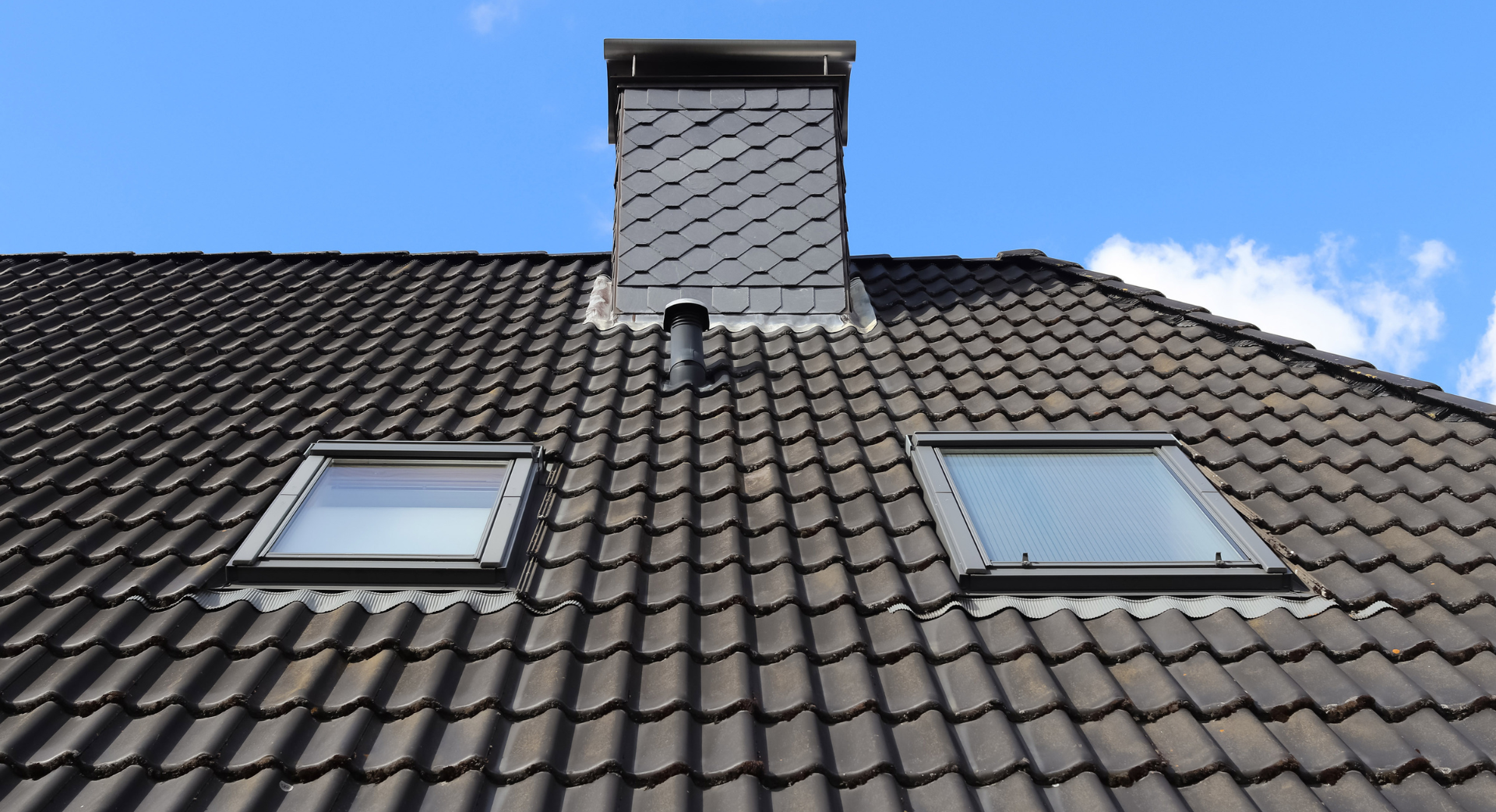 Toit en tuiles gris foncé avec deux puits de lumière et une cheminée sur fond de ciel bleu.