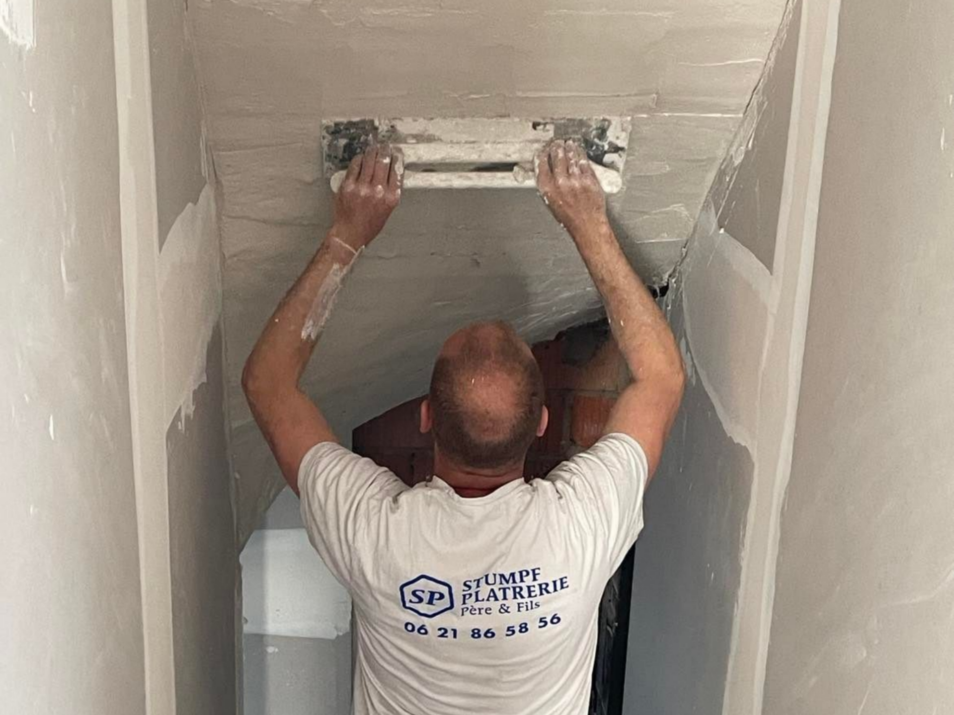 Ouvrier appliquant de l'enduitsur un plafond avec une taloche, t-shirt de l'entreprise