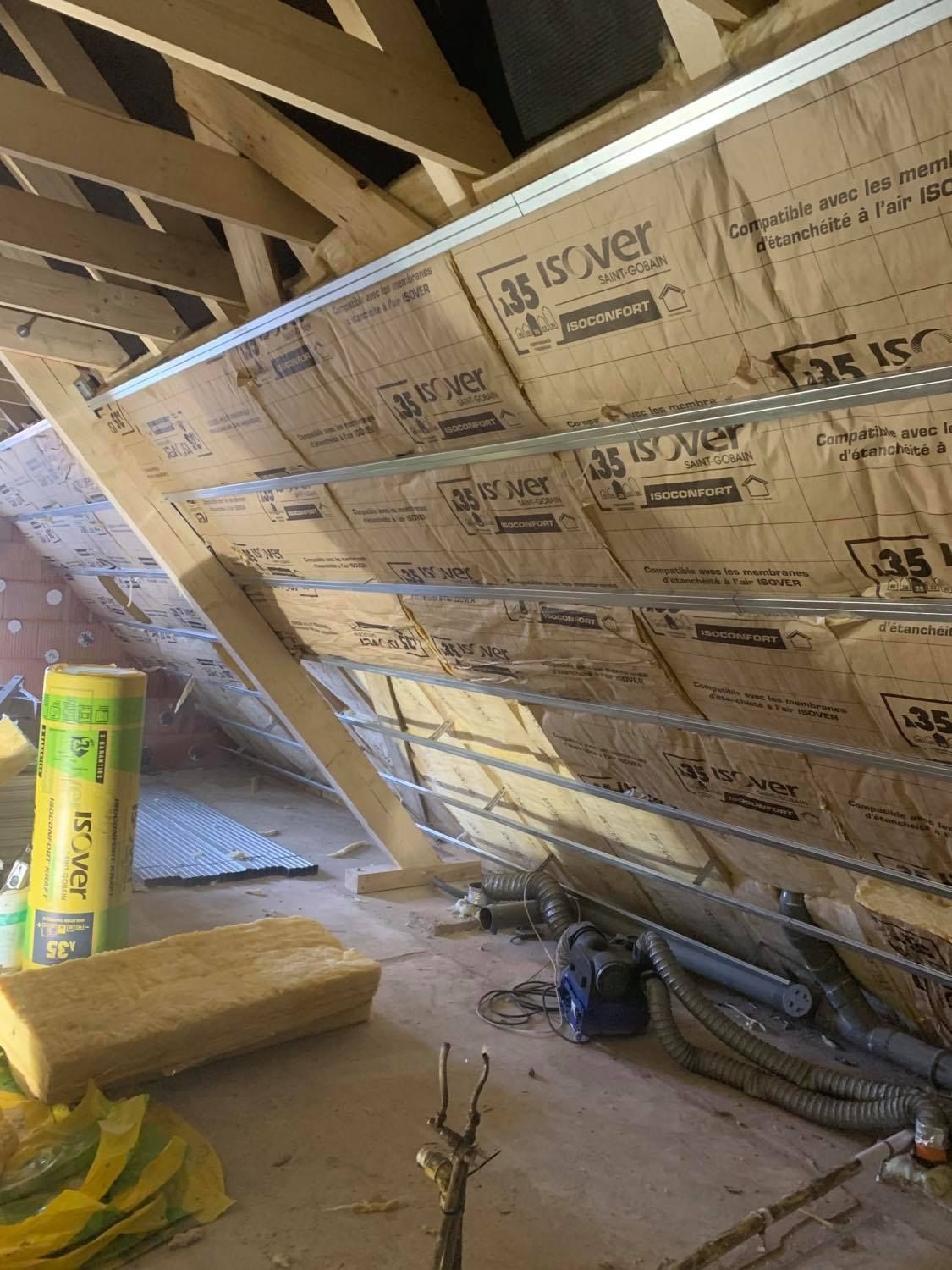 Travaux d'isolation dans des combles de maison, charpente en bois, aspirateur et isolants posés sur le sol