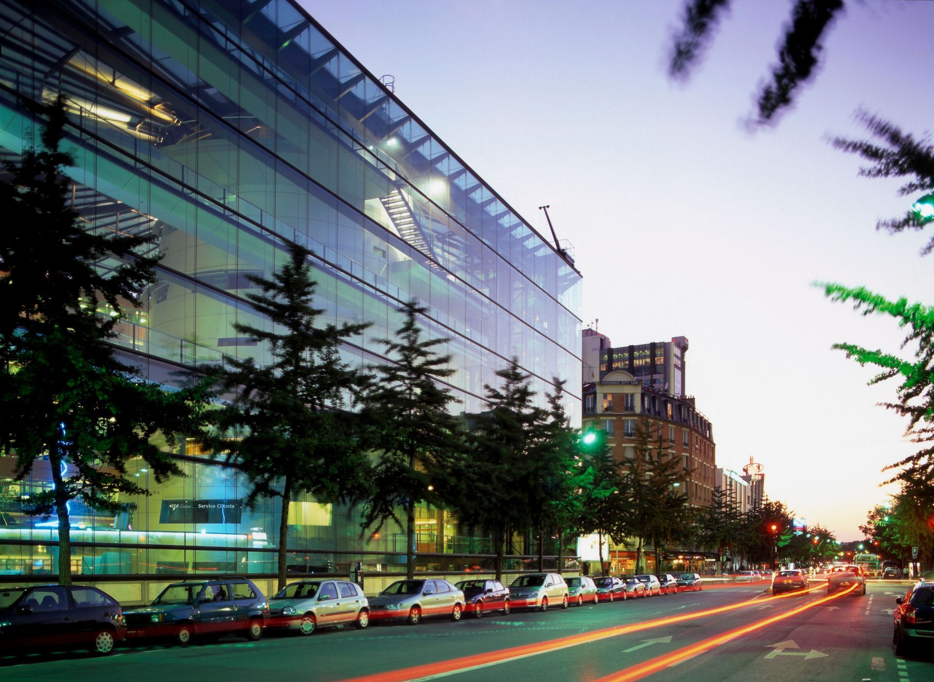 Bâtiment moderne en verre, rue avec des voitures et arbres au crépuscule.