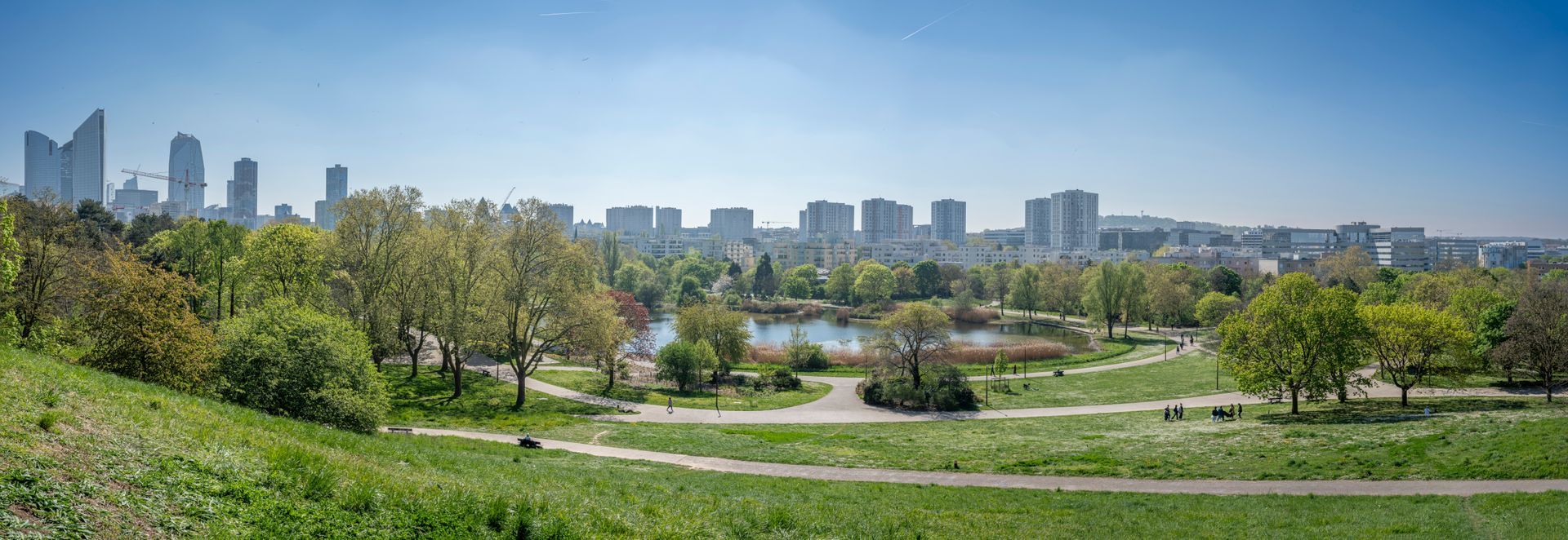 Un parc avec de l'herbe verte et des arbres, et un paysage urbain en arrière-plan sous un ciel bleu.