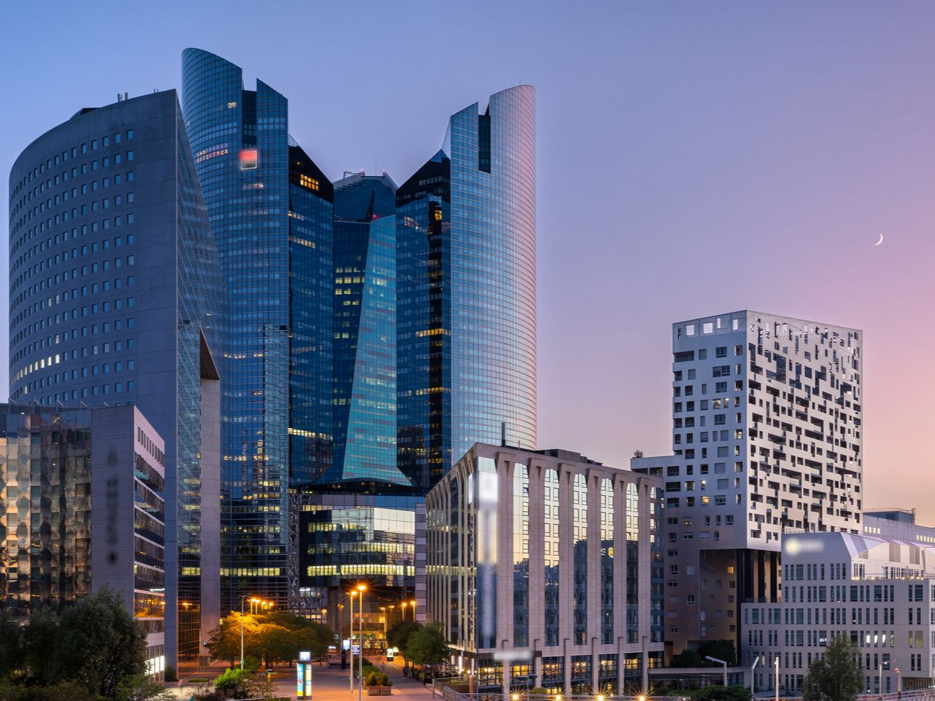 Gratte-ciel d'une ville moderne au crépuscule. Les bâtiments mêlent le bleu, le blanc et le gris, sur un ciel aux teintes pastel.