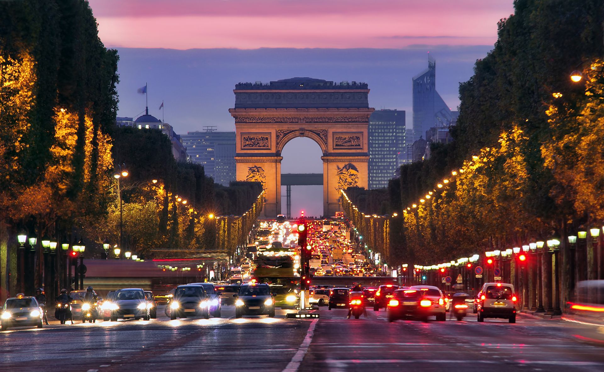 L'Arc de triomphe au crépuscule, vu d'une avenue parisienne bordée de lampadaires et de circulation.