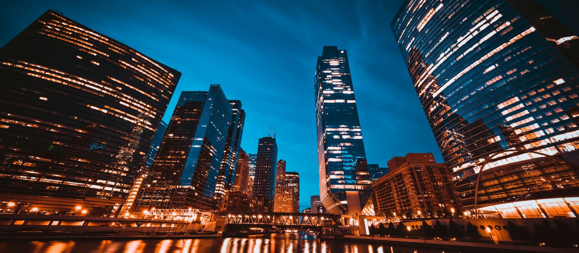 Vue nocturne de la ville, avec ses gratte-ciel illuminés se reflétant dans l'eau.