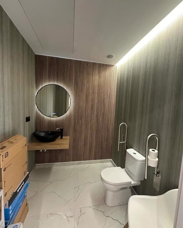 Baño moderno con inodoro, lavabo, espejo redondo, paredes revestidas de madera y suelo de baldosas de mármol.