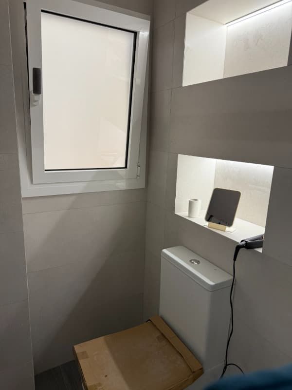 Pequeño rincón de aseo con ventana, estantes en la pared, un armario blanco y un taburete de madera.