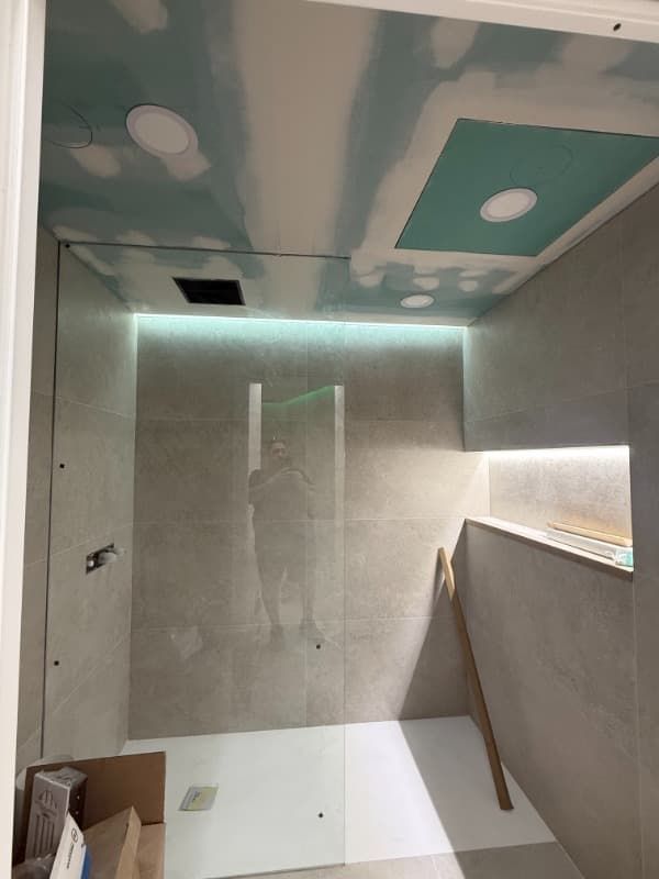 Cabina de ducha parcialmente renovada con azulejos grises, techo sin terminar e iluminación integrada en el estante.