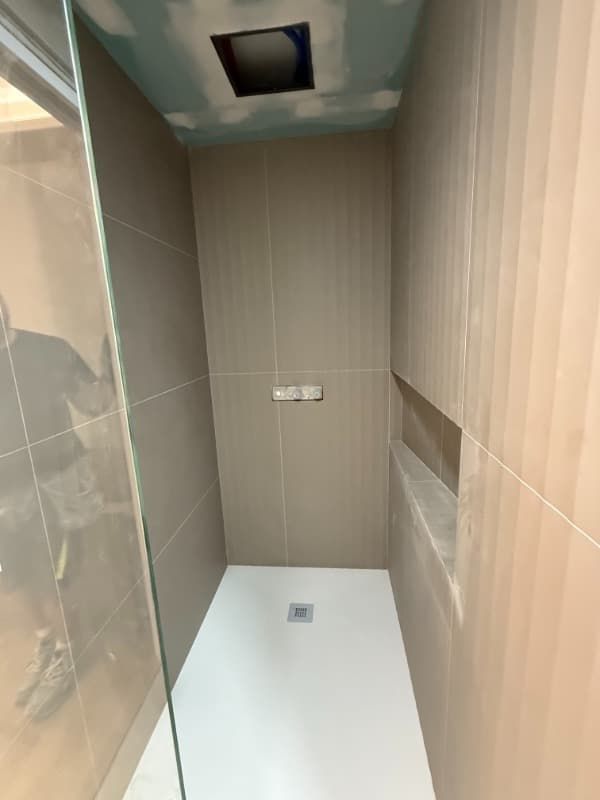 Cabina de ducha estrecha alicatada con suelo blanco y abertura de ventilación en el techo.