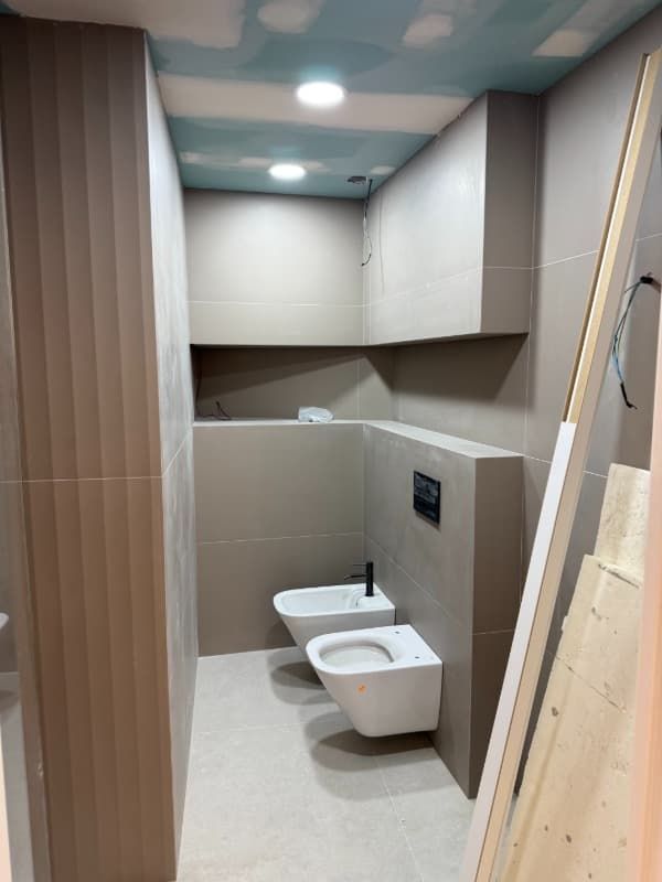 Baño moderno y compacto en construcción con azulejos beige, lavabo mural e inodoro en un espacio estrecho.