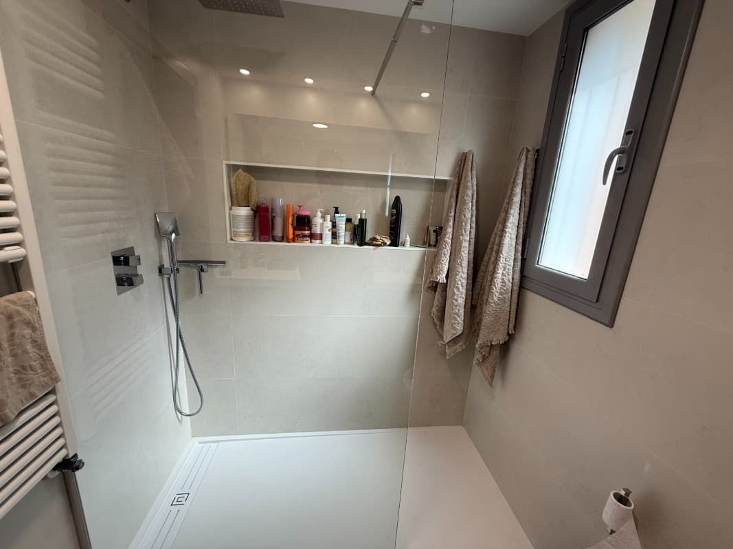 Baño moderno con ducha, azulejos blancos, cabezal de ducha de mano, estante empotrado y toallas colgadas junto a la ventana.