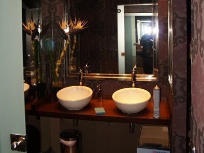 Tocador de baño moderno con dos lavabos blancos sobre encimera, espejo, flores e iluminación cálida.
