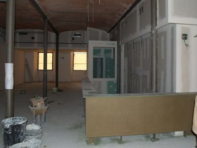Interior en construcción con estructura expuesta, paneles de yeso y materiales de pintura sobre un suelo polvoriento.