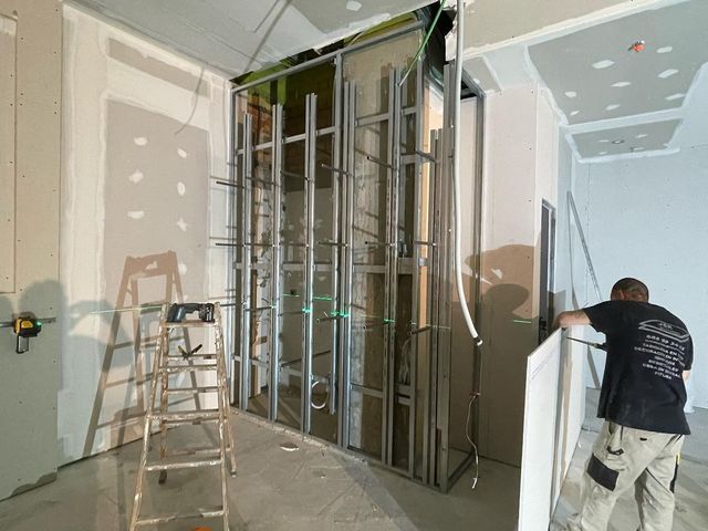 Obra de construcción de paneles de yeso con montantes metálicos, escalera y un trabajador instalando un marco de pared.