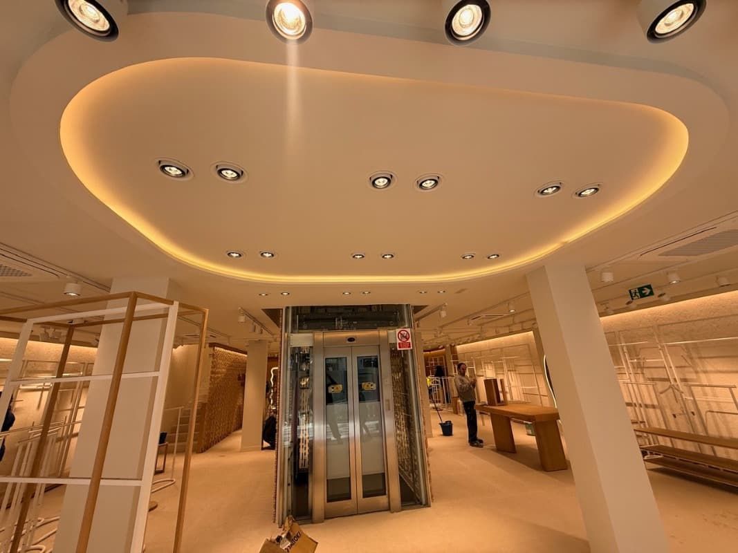 Interior de tienda minorista moderna con iluminación cálida en el techo, ascensor de cristal y vitrinas para ropa.