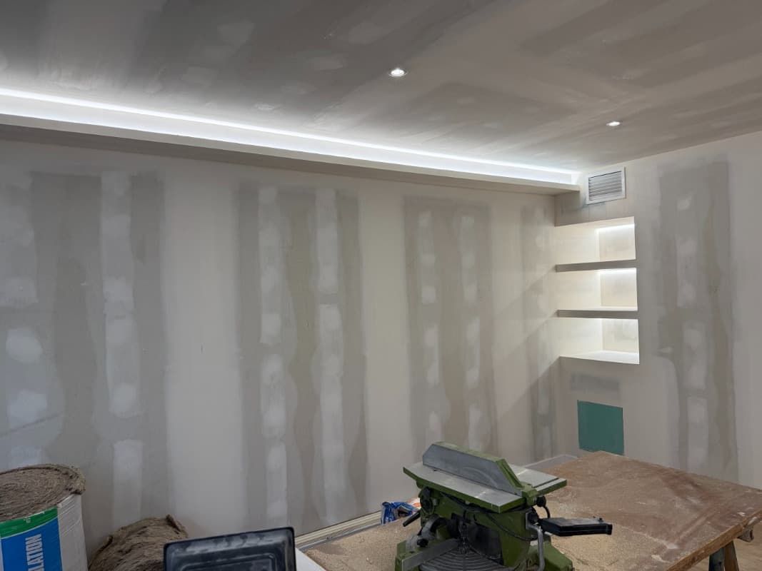 Habitación parcialmente pintada con paredes blancas, luces en el techo y un banco de trabajo cubierto de herramientas y materiales.