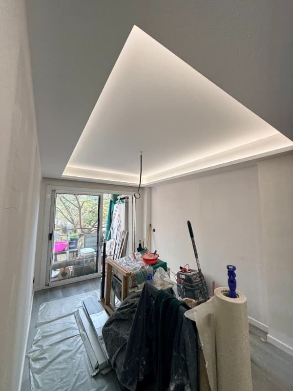 Cuarto de reformas con techo triangular iluminado, suelo cubierto, herramientas y puerta de acceso a un balcón.
