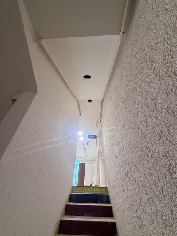 Estrecha escalera interior con paredes blancas, pequeñas luces en el techo y una puerta luminosa en la parte superior.