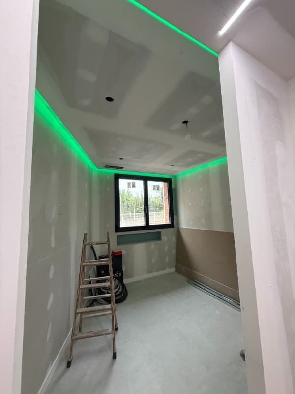 Habitación interior en proceso de renovación con luces LED verdes en el techo, una escalera y una ventana.
