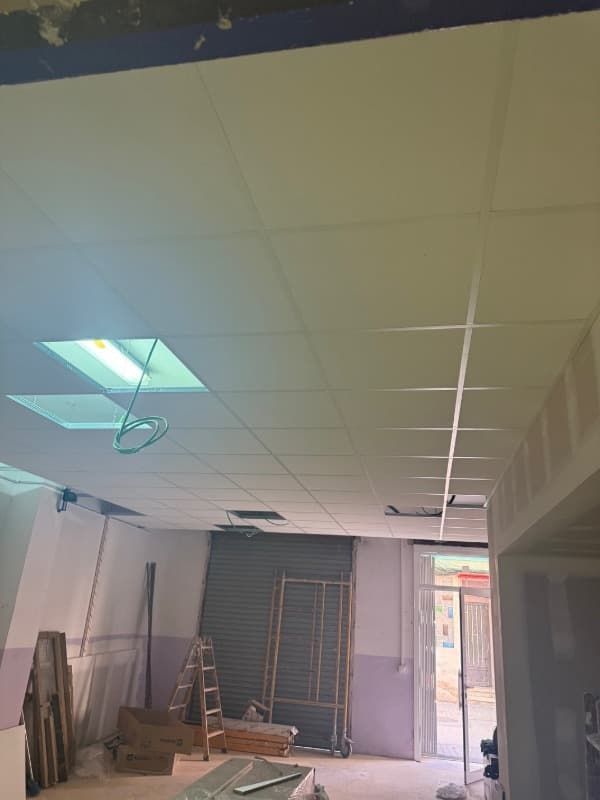 Interior en proceso de renovación con paneles de techo blancos, escalera, herramientas y una puerta luminosa en la parte trasera.