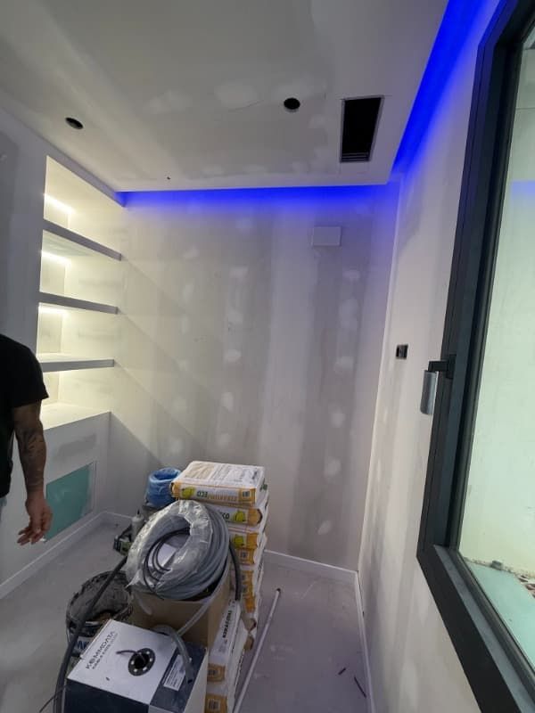 Habitación pequeña sin terminar con luces LED azules en el techo, materiales apilados y una ventana a la derecha.