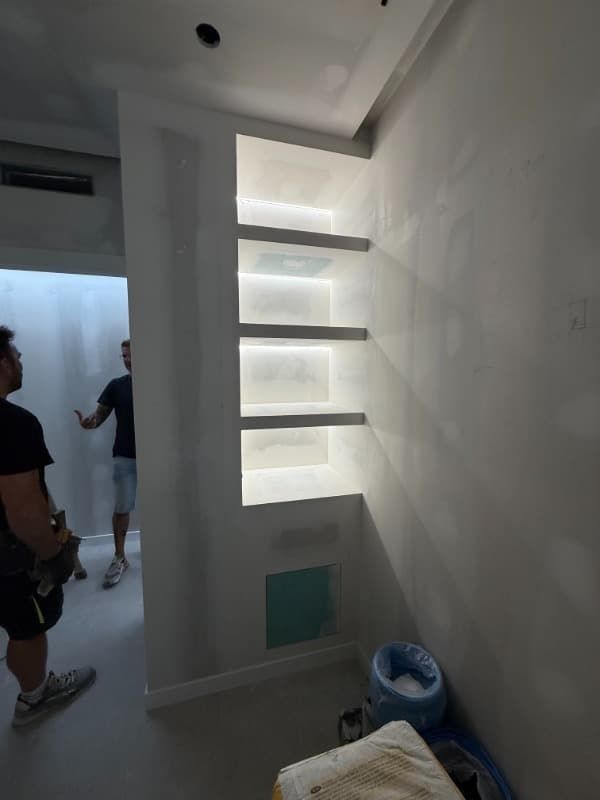 Un estrecho pasillo blanco con estanterías iluminadas integradas y dos personas cerca de una puerta.