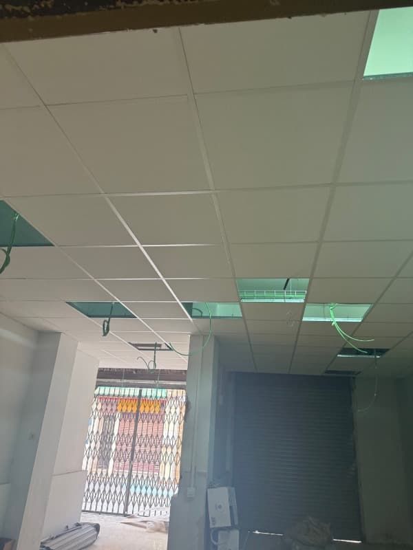 Interior de oficina con falso techo de baldosas, luces fluorescentes, puerta abierta y ventana con barrotes.