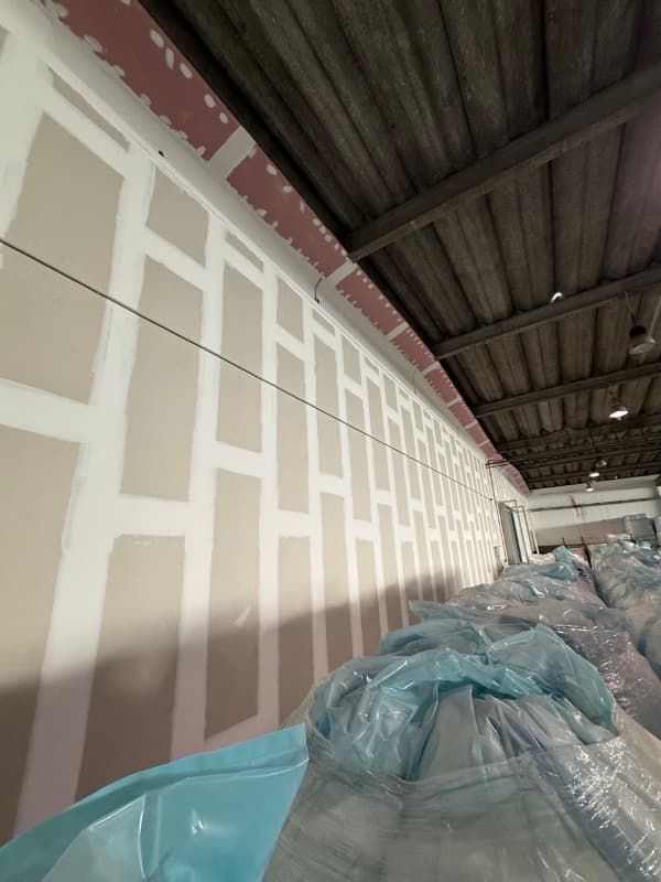 Pared de almacén parcialmente pintada bajo un techo de chapa ondulada, con los materiales cubiertos con láminas de plástico.