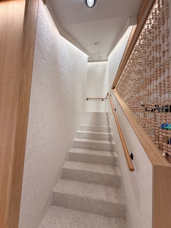 Escalera interior estrecha con paredes blancas texturizadas, escalones alfombrados de color claro y pasamanos de madera.