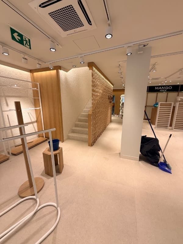 Interior de una tienda con escaleras, un pilar blanco, una escalera y productos de limpieza en el suelo.