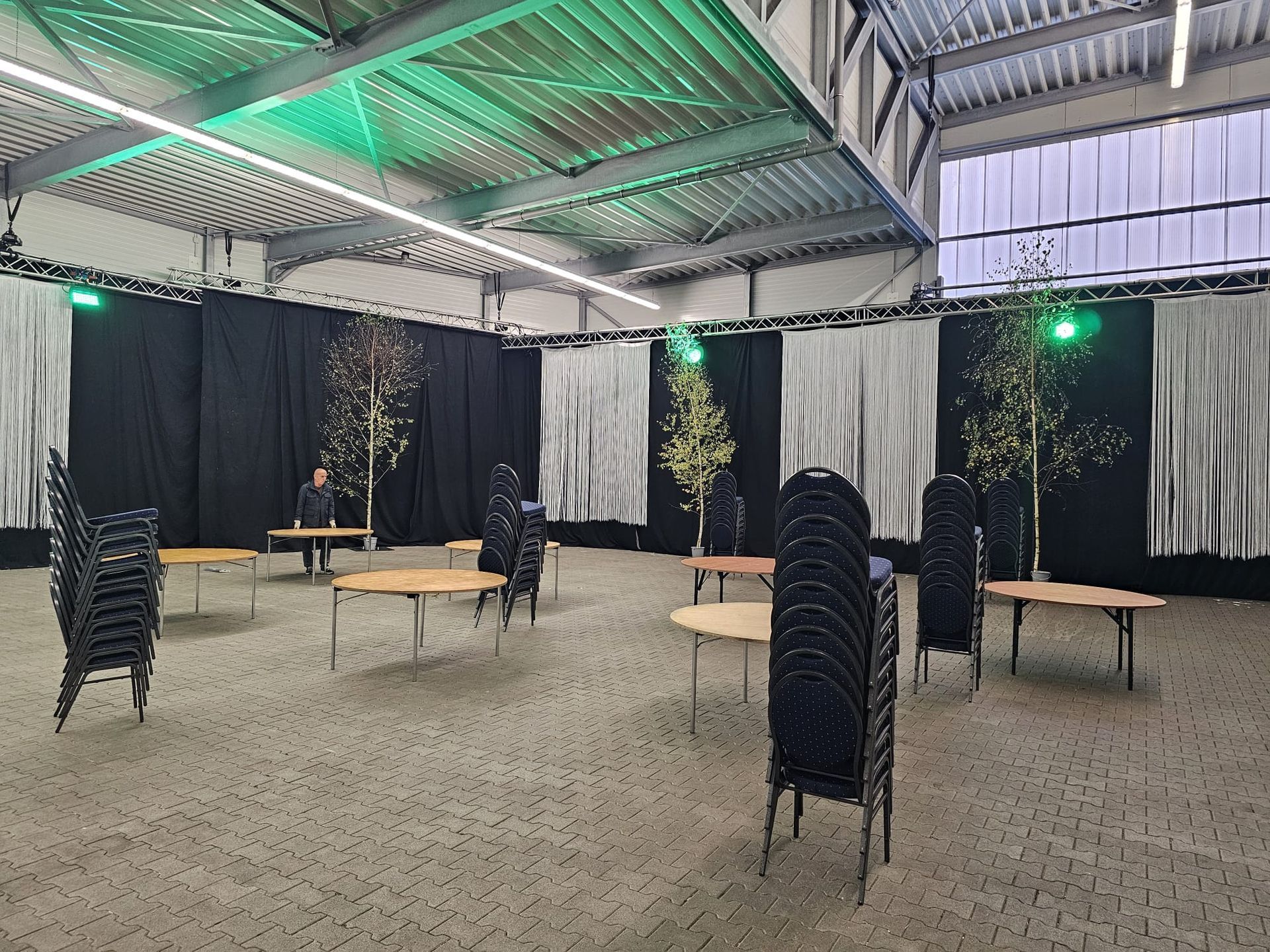 Ein Veranstaltungsraum mit runden Tischen, Stühlen und Bäumen vor schwarz-weißen Vorhängen und grüner Deckenbeleuchtung.