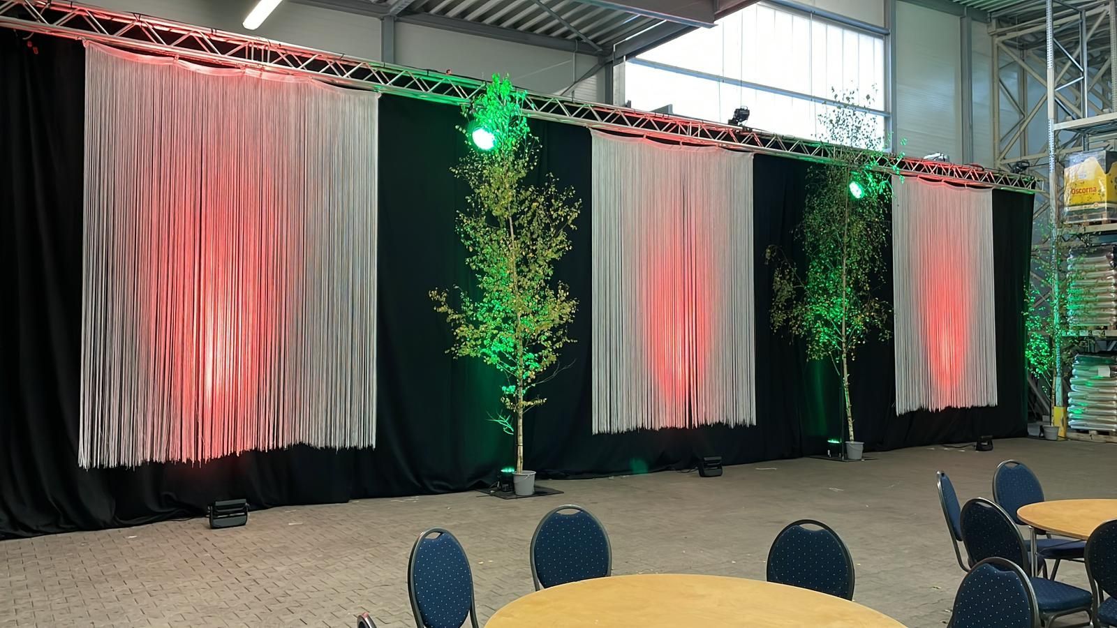 Hintergrund der Veranstaltung: schwarz, weiß und rot drapierte Tafeln mit Bäumen, beleuchtet von grünen und roten Lichtern. Tische und Stühle im Vordergrund.