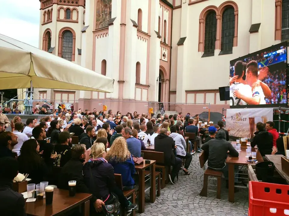 Eine große Gruppe von Menschen sitzt vor einer Kirche und schaut die Fußball-WM Liveübertragung