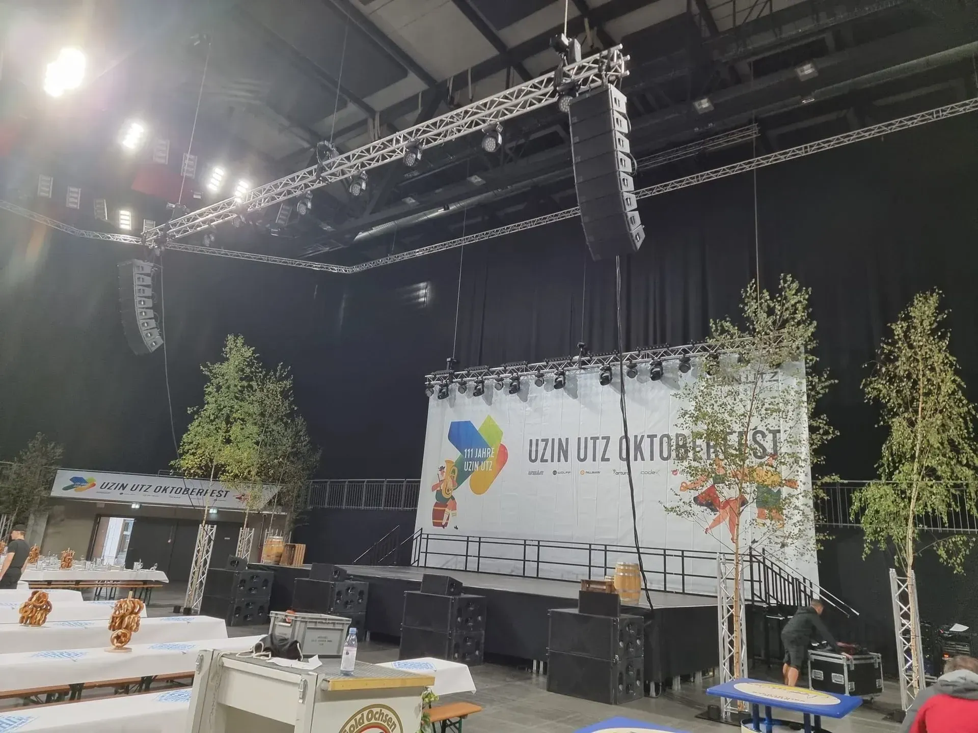 Oktoberfest in einer Eventhallt mit Bühne, Bäumen und Bierzeltgarnitur