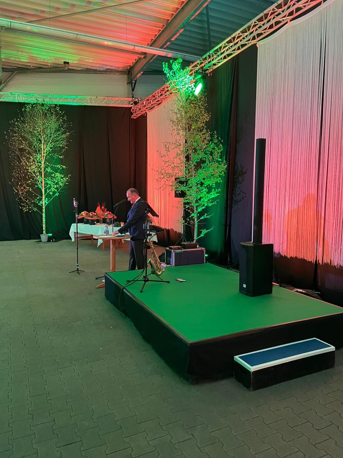 Mann spielt Saxophon auf einer Bühne mit grünem Teppich. Birken und Beleuchtung schmücken den Veranstaltungsort.