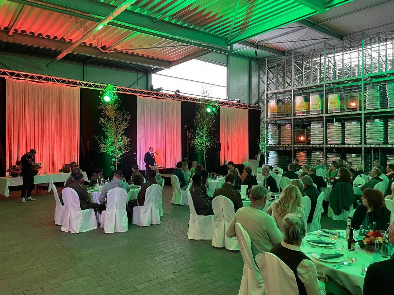 Veranstaltung in einer Lagerhalle: Menschen sitzen an Tischen, Bühne mit Lautsprechern, grüne und rote Beleuchtung.
